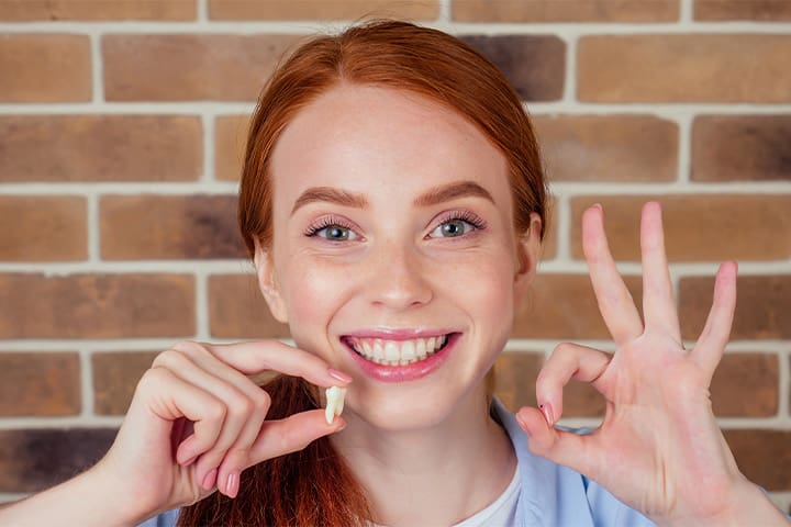 A smiling, red-headed girl holding an extracted tooth and giving an okay sign